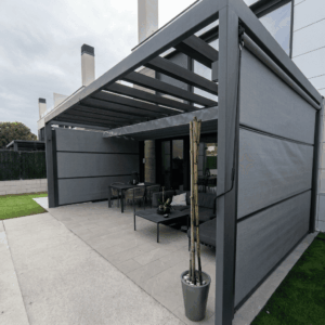 Una pérgola moderna de aluminio gris oscuro instalada en una terraza adoquinada con un comedor exterior y un sofá rinconera. Los paneles laterales retráctiles de tela gris están parcialmente bajados, mientras que el techo de lamas de aluminio está abierto. Una maceta con bambú está en primer plano. El cielo está nublado y se ven casas adosadas modernas al fondo.
