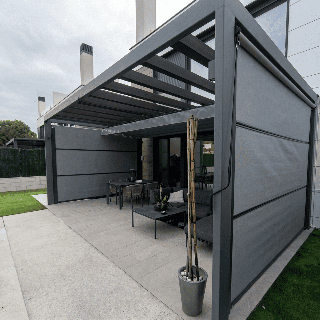 Una pérgola moderna de aluminio gris oscuro instalada en una terraza adoquinada con un comedor exterior y un sofá rinconera. Los paneles laterales retráctiles de tela gris están parcialmente bajados, mientras que el techo de lamas de aluminio está abierto. Una maceta con bambú está en primer plano. El cielo está nublado y se ven casas adosadas modernas al fondo.