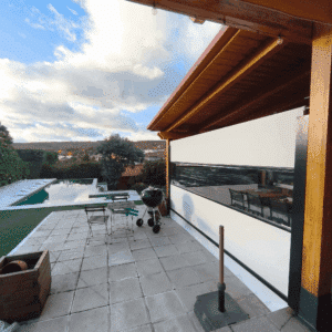 Porche con techo de madera y toldo cortavientos vertical blanco con ventana transparente, situado en un patio con barbacoa y jardín con piscina al fondo.
