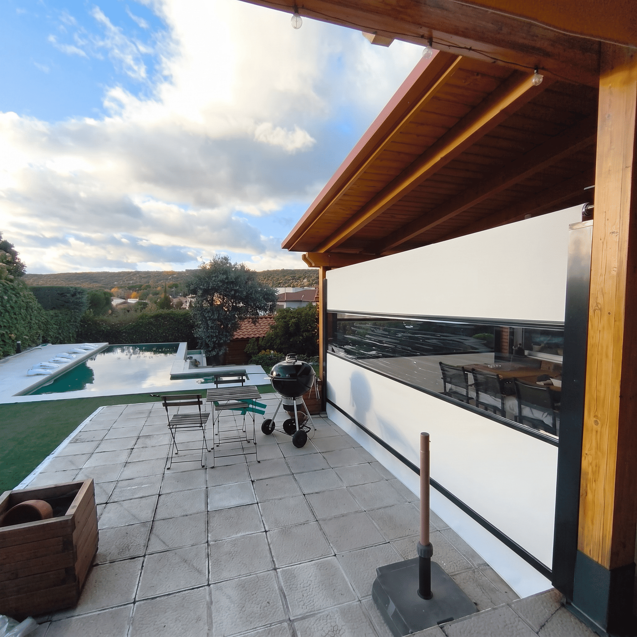 Porche con techo de madera y toldo cortavientos vertical blanco con ventana transparente, situado en un patio con barbacoa y jardín con piscina al fondo.