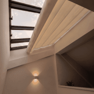Lucernario interior con estructura oscura y toldo corredero de tela beige parcialmente extendido en una habitación de paredes blancas con luz cálida.