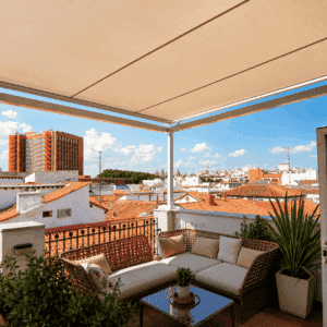 Terraza de ático amueblada con sofá de ratán, plantas y vistas panorámicas de los tejados tradicionales de Sevilla bajo un toldo extendido.