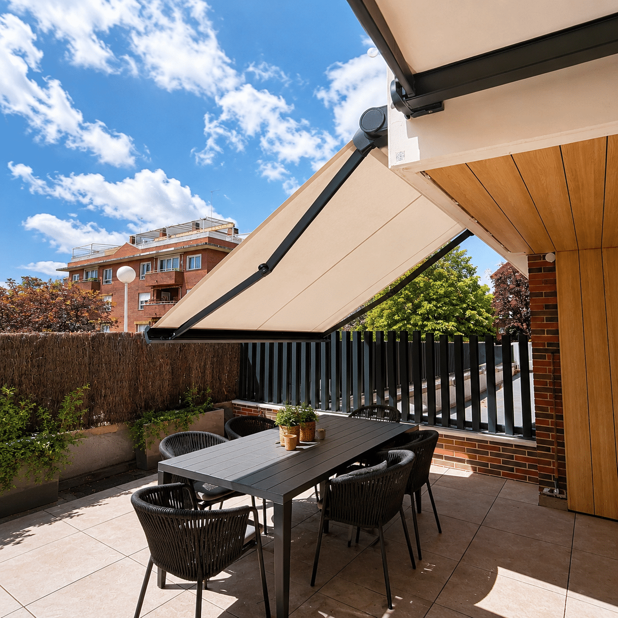 Toldo cofre de brazos articulados oscuros y lona beige extendido sobre un comedor de exterior en una terraza soleada.